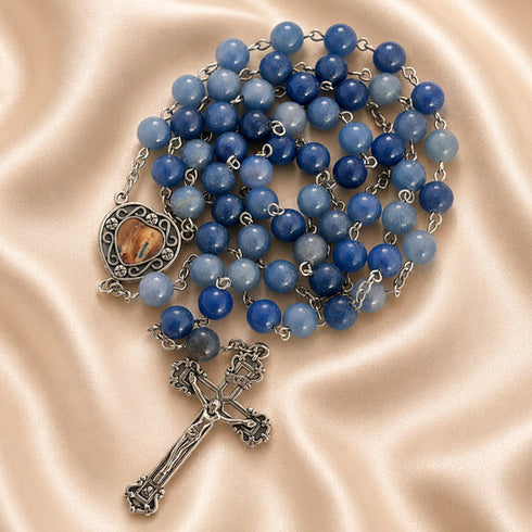 A blue stone rosary rosary with a silver-toned crucifix and heart-shaped medallion, on a beige satin fabric background.