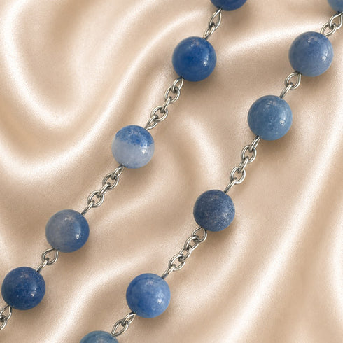 A beaded necklace featuring blue gemstone beads linked by silver-toned metal chains, on a beige fabric background.
