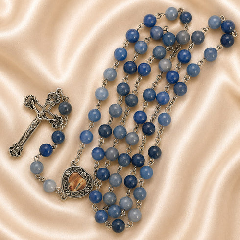 A rosary necklace with blue beads, a silver-tone crucifix, and a heart medallion, on a beige satin fabric background.