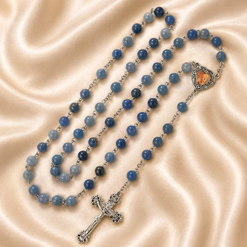 A rosary with light and dark blue beads, a silver crucifix, and a heart-shaped medallion, on a beige satin fabric.