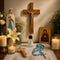 A religious tabletop featuring a wooden crucifix on the wall, a Virgin Mary statue, a Holy Water bottle, blue bead rosary, candles.