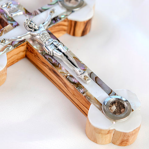 Detail of the bottom of an olive wood budded cross with holy soil capsule adorned with a silver crucifix and a white and green mother of pearl, resting on a clean white surface.