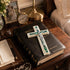 Decorative cross on a Holy Bible with a pen and photo frame in the background.
