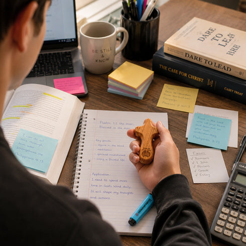Person holding a comfort cross, studying at a desk with books, laptop, and stationery