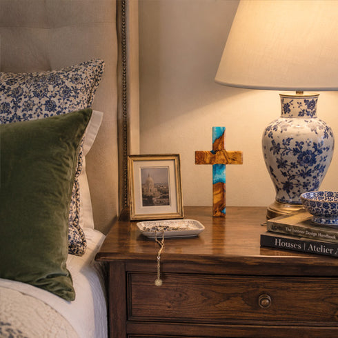 Nighstand with decorative items including a lamp, cross, and books next to a bed with patterned pillows.