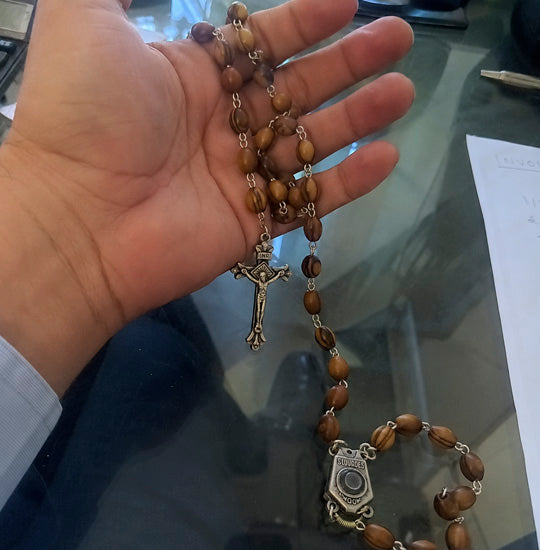 Hand holding a wooden rosary with a cross and medal on a reflective surface.