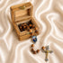 A wooden rosary and a gold- and silver-crucifix, displayed beside an open wooden box on draped satin fabric.