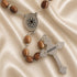 A wooden and silver rosary, a round silver centerpiece with a Jerusalem cross laid out on a satin fabric background.