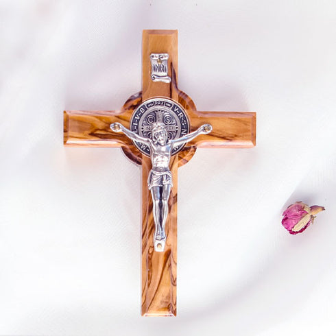 Frontal image of a handcrafted olive wood cross with St. Benedict medallion and silver-toned crucifix detail on a white background with petals.
