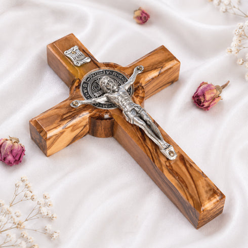 Handcrafted olive wood cross with St. Benedict medallion and silver-toned crucifix detail on a white background with petals.