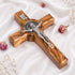 Handcrafted olive wood cross with St. Benedict medallion and silver-toned crucifix detail on a white background with petals.