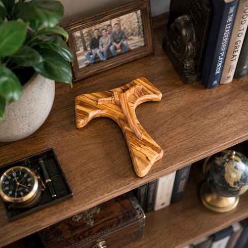 Wooden Tau cross on a wooden shelf with books, a clock, and a photo frame.