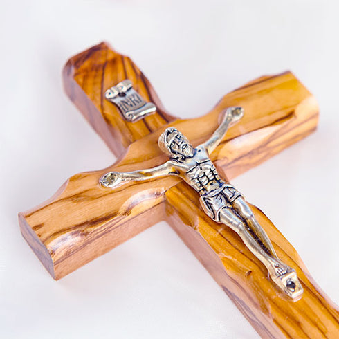 A handmade olive wood thorn cross with a crucifix, placed on a white surface with a few pink flowers scattered around.
