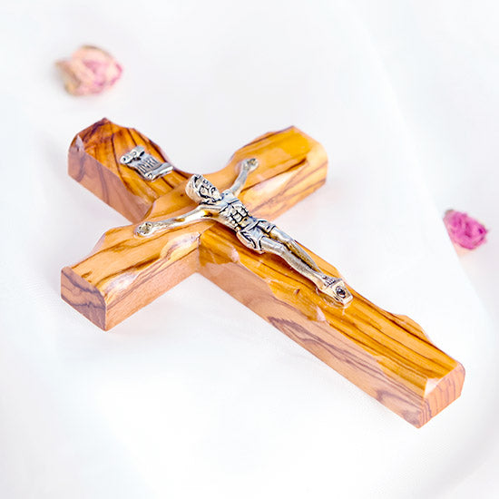 A handcrafted olive wood thorn cross with a crucifix, placed on a white surface with a few pink flowers scattered around.