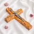 A handcrafted olive wood thorn cross with a crucifix, placed on a white surface with a few pink flowers scattered around.