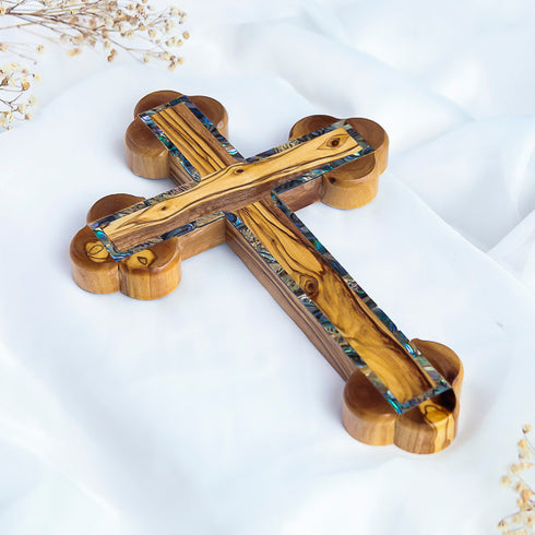 A handcrafted olive wood budded cross with green mother-of-pearl inlay, featuring the Twelve Stations of the Cross engraved on the back, placed on a white fabric surface.