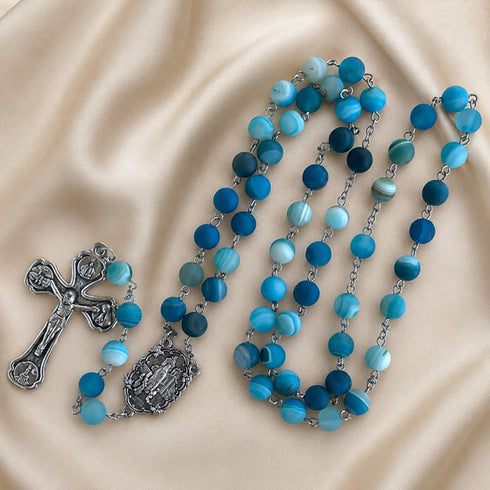 A rosary with blue and turquoise beads on a silver chain, a silver crucifix, on a beige satin fabric background.