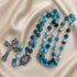 A rosary with blue and turquoise beads on a silver chain, a silver crucifix, on a beige satin fabric background.