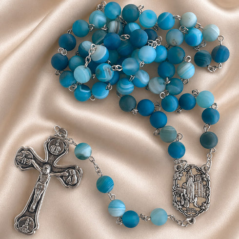 A blue beaded rosary with silver-tone metal links, a crucifix, and a medallion, on draped satin fabric.