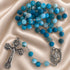 A blue beaded rosary with silver-tone metal links, a crucifix, and a medallion, on draped satin fabric.