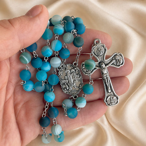 A close-up of a rosary held in a person's hand, featuring turquoise and light blue marbled beads.