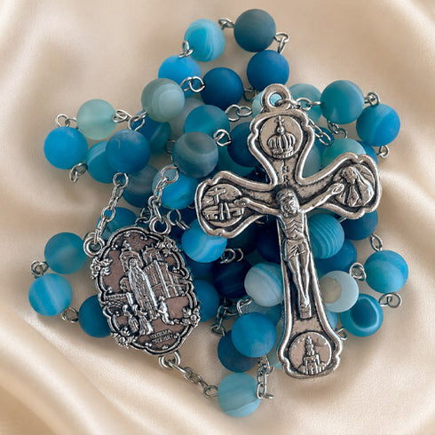 A blue and turquoise rosary with metal crucifix and medallion, on a cream satin fabric background.