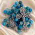 A blue and turquoise rosary with metal crucifix and medallion, on a cream satin fabric background.