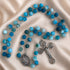 A blue rosary with silver-tone metal links, a crucifix and an oval medallion, on a beige satin background.