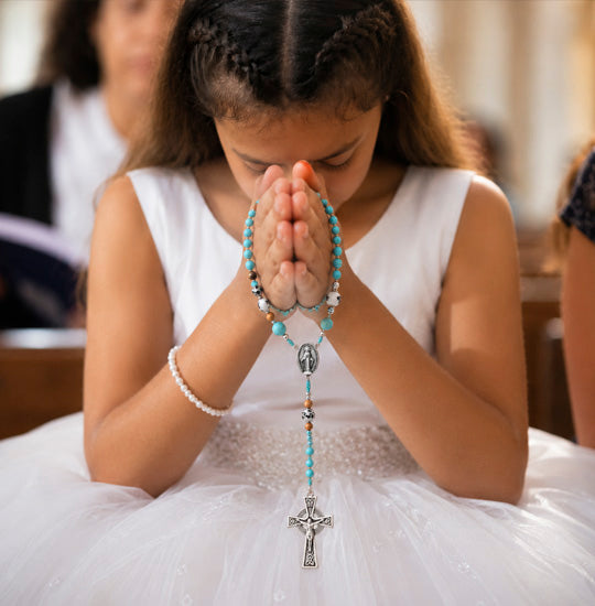 Young girl in a white dress holding a rosary with her hands clasped together.