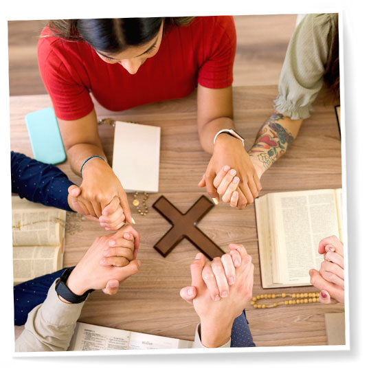 Bird view and close up of people holding hands and praying over a wooden cross on a table.