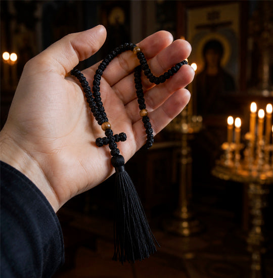 Hand holding a black rosary with a blurred background of candles and religious artwork