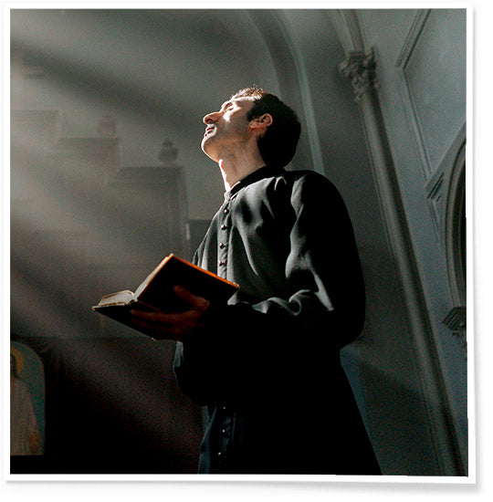 Priest dressed in black holding a brown Bible, looking up and praying in a stone church.