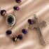 A close-up of a rosary necklace with faceted purple and blue beads and a silver-tone cross, on a satin beige fabric background.