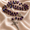 A rosary with purple beads, silver-tone metal links, a silver medallion, and a silver crucifix, displayed on draped satin fabric.