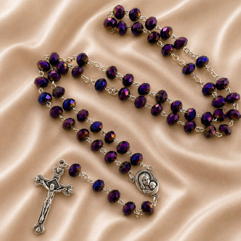 A rosary with purple beads on a silver chain, a silver crucifix and a silver centerpiece, on a beige satin fabric background.