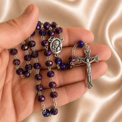A rosary with purple beads and silver-tone metal links, held in a person's hand against a satin beige fabric background.
