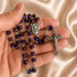 A rosary with purple beads and silver-tone metal links, held in a person's hand against a satin beige fabric background.