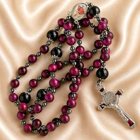 A gothic-style rosary with red and black beads, a silver-toned crucifix and a medallion, on a satin fabric.