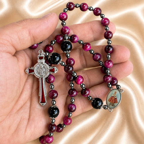 A close-up of a rosary draped over a hand, with red and black beads, a silver-toned crucifix, oval saint medal, on a beige background.