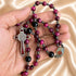 A close-up of a rosary draped over a hand, with red and black beads, a silver-toned crucifix, oval saint medal, on a beige background.