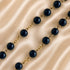 A close-up of a beaded chain necklace with glossy dark faceted beads separated by gold-tone links, laid on a satine background.