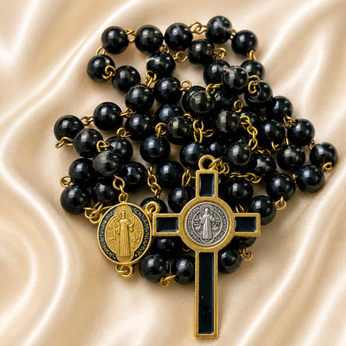 Black and gold rosary with polished black beads, a gold cross pendant, and a round medallion, on a cream satin fabric background.