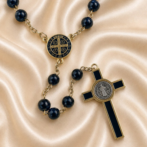 A Catholic rosary with dark beads, a Saint Benedict medallion, and a cross, on a draped beige satin background.