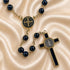 A Catholic rosary with dark beads, a Saint Benedict medallion, and a cross, on a draped beige satin background.