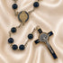 A Catholic rosary with black glass beads and a St Benedict cross, on a draped beige satin fabric background.