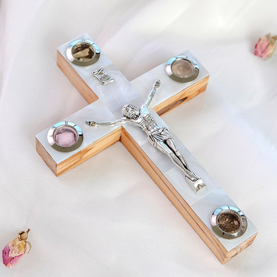 Detailed image of a small classic olive wood Latin cross featuring silver crucifix, white Mother of Pearl inlay and Four Sacred Elements capsules, with soft rose petals around it on a clean white surface.