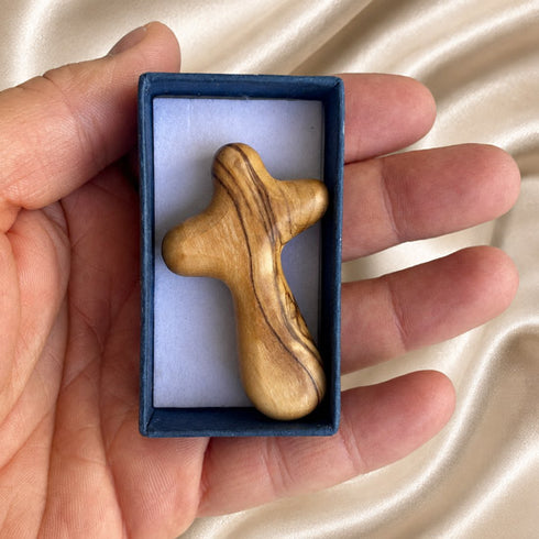 A hand holds a small carved wooden cross inside an open dark blue presentation box.
