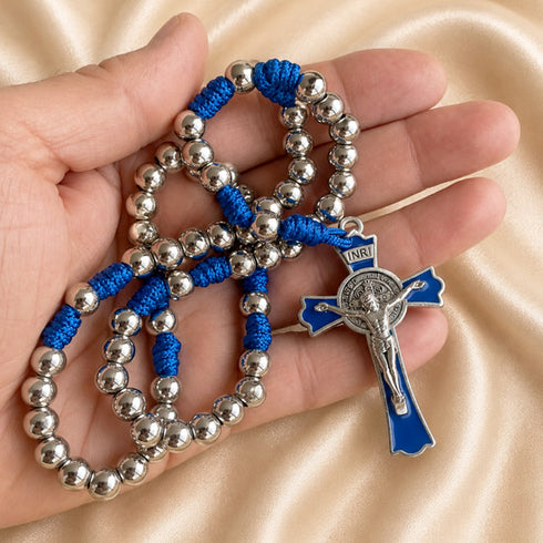 A close-up of a rosary with metal beads and a blue-and-silver crucifix pendant, held in a person's hand on a satin beige fabric.