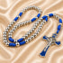 A rosary with metal beads and a matching crucifix pendant, accented with blue braided cord, on a beige background.