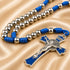 A close-up of a rosary with blue cord and metal beads, a blue and silver crucifix, on a beige satin fabric background.